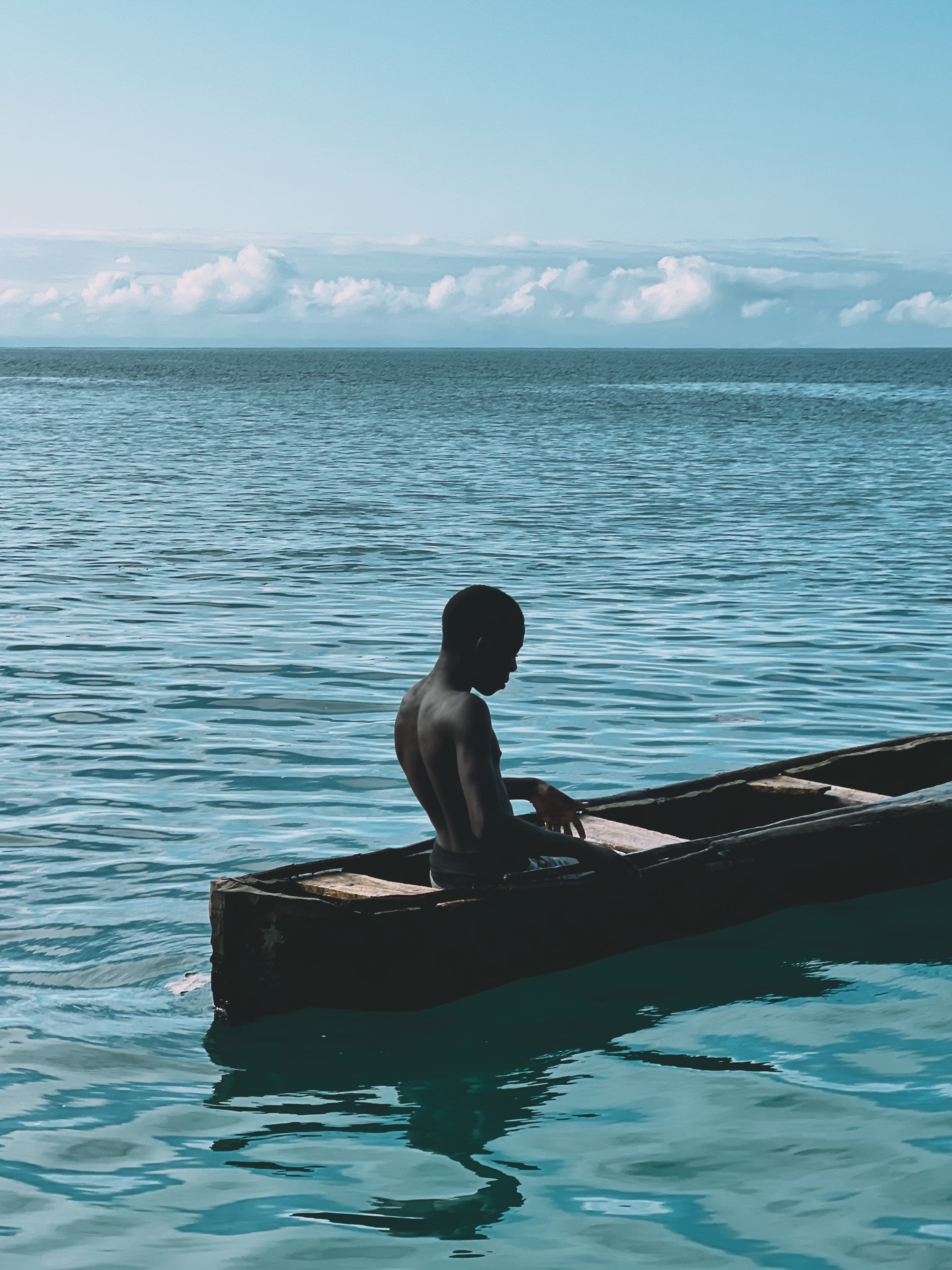 Um jovem sentado sem camisa em um pequeno barco a remo alongado no mar, olhando para longe, visto de um ângulo traseiro diagonal
