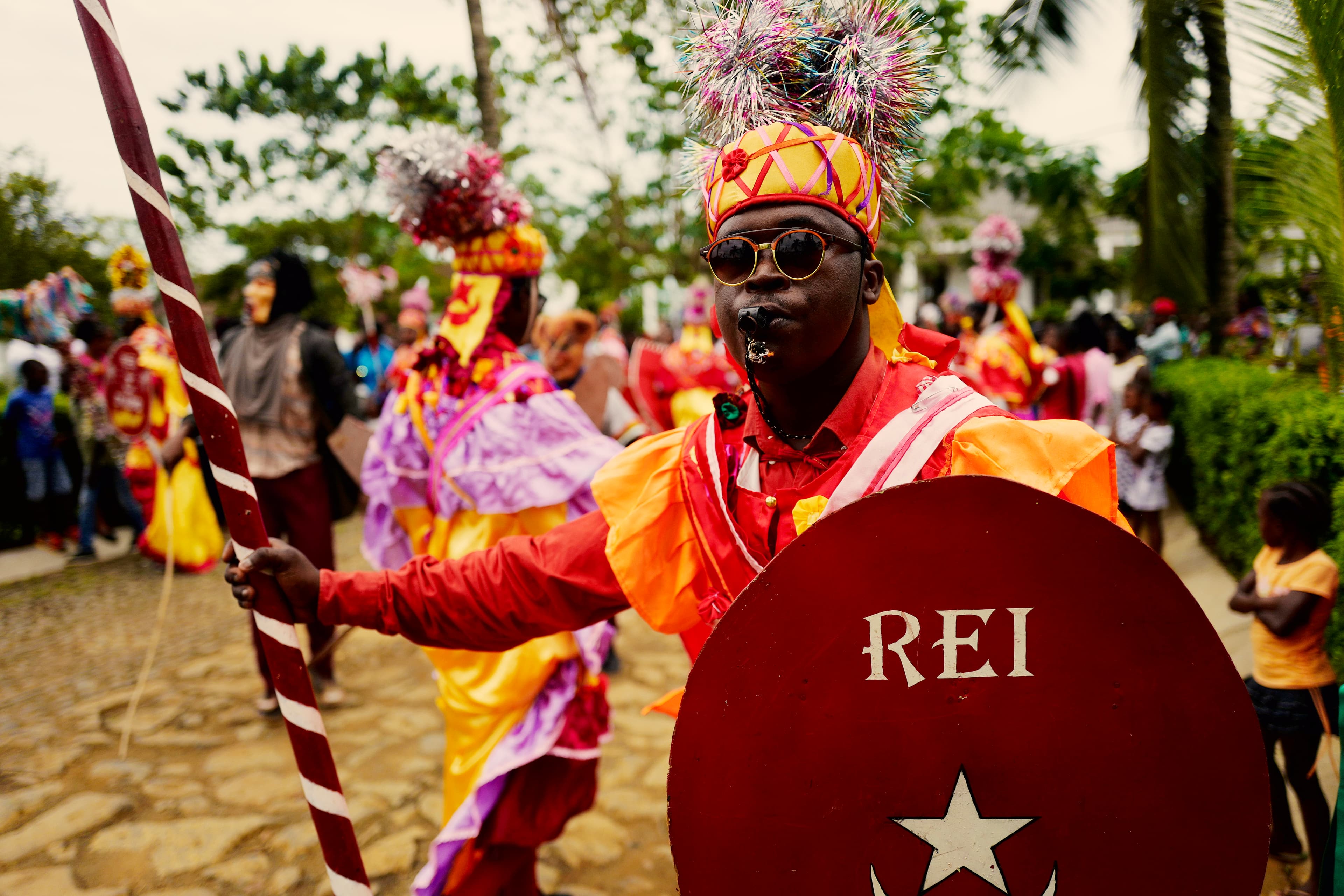Um homem com traje tradicional em tons vibrantes de laranja e vermelho, com um elaborado adereço de cabeça