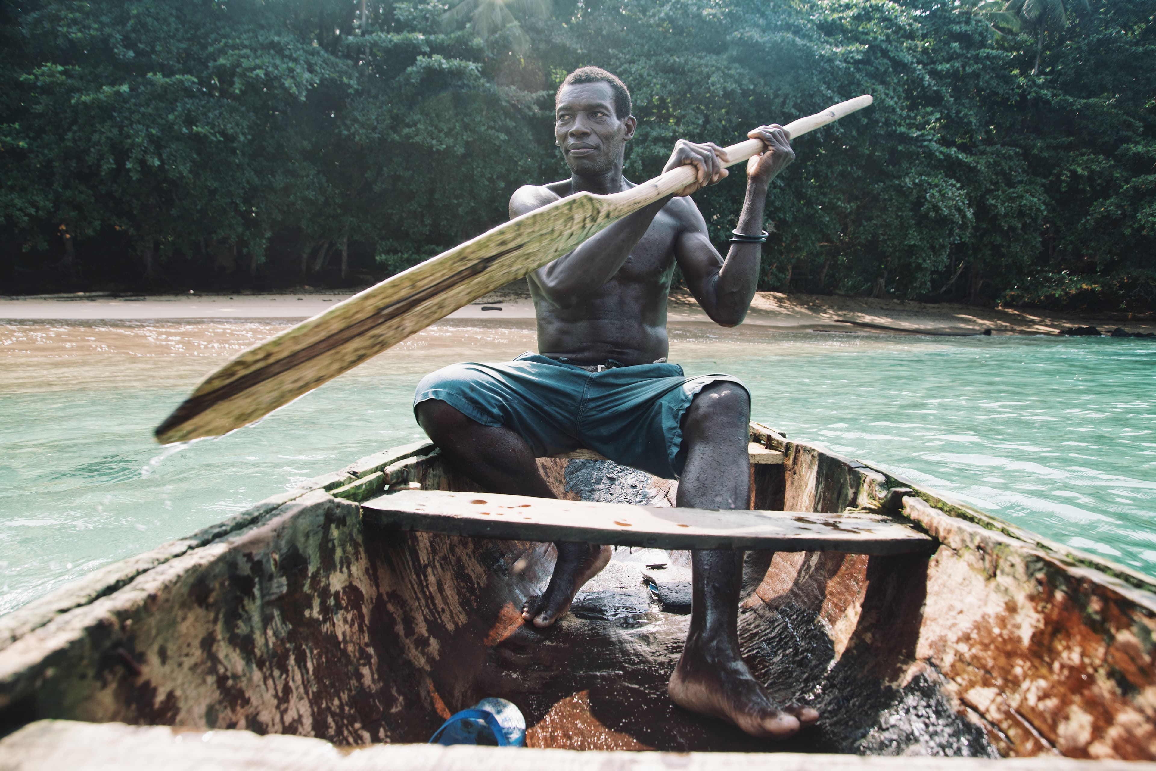 Homem sentado num barco tradicional de madeira com calções de banho azul escuro, a remar sobre água turquesa com um remo de madeira, ilha ao fundo